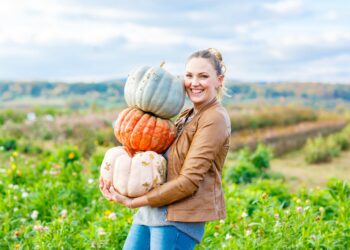 Junge, lachende Frau steht auf einem Feld und hält drei unterschiedlich geformte Kürbisse.