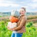 Junge, lachende Frau steht auf einem Feld und hält drei unterschiedlich geformte Kürbisse.