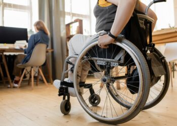 Eine Frau im Rollstuhl bewegt sich auf eine Kollegin zu, die an einem Tisch sitzt. Die Szene spielt sich in einem modernen, barrierefreien Innenraum ab und zeigt gelebte Inklusion am Arbeitsplatz.