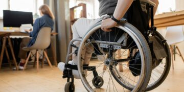 Eine Frau im Rollstuhl bewegt sich auf eine Kollegin zu, die an einem Tisch sitzt. Die Szene spielt sich in einem modernen, barrierefreien Innenraum ab und zeigt gelebte Inklusion am Arbeitsplatz.