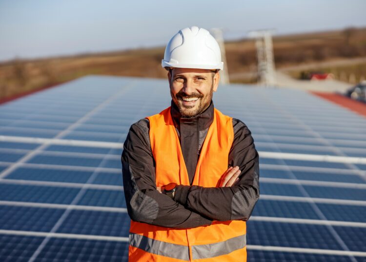 Das Symbolbild zeigt einen lächelnden Handwerker, im Hintergrund sind Sonnenkollektoren zu sehen.